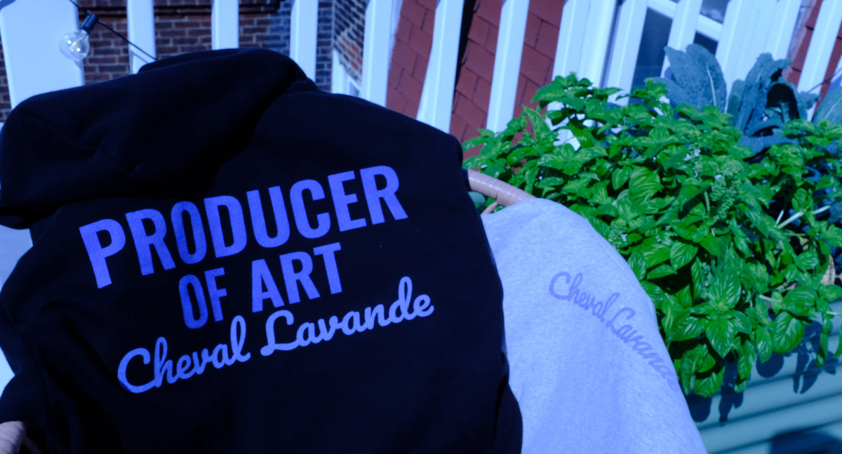 A black hoodie with blue and white text reading 'PRODUCER OF ART Cheval Lavande' draped over a light gray sweatshirt in an outdoor setting with a potted plant in the background.