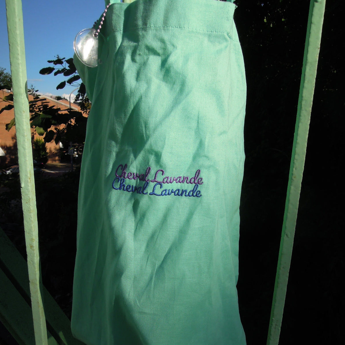 A mint green fabric bag with purple and blue embroidered text reading 'Cheval Lavande' hanging outdoors on a green metal bride. 